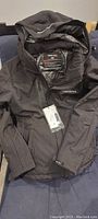 Full view of the black Venustas heated jacket showing hood, full zipper, and tags.
