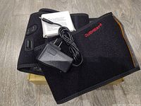 Black CueHeat branded cordless heating pad laid flat on floor with visible control buttons, battery pocket, power adapter with plug and cable, and instruction manual above it.