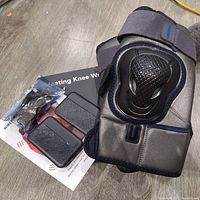 Photo showing the Ginekoo heated knee massager device, charging accessories, and instruction manual laid on a wood floor.