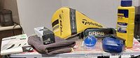 Photo of golf accessories including Taylormade headcovers, golf balls, gloves, towels, blue ball washers, and a yellow chalk line canister