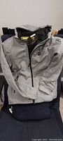 Front view of grey Ptahdus heated jacket laid out on chair, showing hood, zipper, and logo on sleeve.