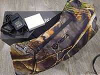 Savior Heat Heated Hand Muff with camouflage pattern, black zipper pocket with logo, an AC adapter charger, and a small white box on wooden floor.