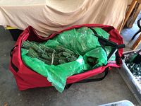 Red canvas bag open to show wrapped artificial tree inside with metal stand included
