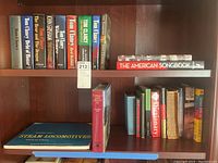 Two wooden shelves containing hardcover books. Top shelf includes Tom Clancy titles, American Songbook, and Steam Locomotives. Bottom shelf has more hardcover books including a dictionary and smaller books.