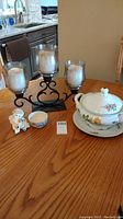 Overview of lot items on wooden table including soup tureen, triple candle holder, Dreamsicles figurine, and small bowl.