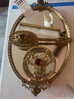 Oval vintage vanity mirror with detailed gold-tone filigree frame, amber-colored decorative stones, matching ornamental hair comb and brush.