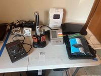 Photo showing two Midland walkie talkies in charging dock, a flashlight, an analog clock, a digital alarm clock, VOIP phone handset in packaging, and multiple small electronics items on white table.