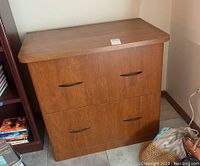 Photo of composite wood file cabinet showing front with two large drawers and black handles in a medium brown finish, positioned next to a bookcase and other household items.