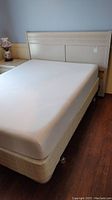 Full image showing the headboard attached to a mattress and box spring, cream color, positioned against a wall with nightstand partially visible.