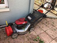 Full side view of Honda gas powered lawn mower and red gas can side by side on tiled outdoor surface with leaves scattered.