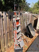Full image of the Little Giant aluminum ladder with ratchet and levelers leaning against a wooden fence with a black wheelbarrow nearby.