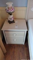 Front and top view of cream-colored nightstand with lamp on top showing overall condition and design.