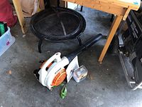 Photo showing black metal fire pit with mesh cover and four legs, and the leaf blower with owner's manual and fuel can