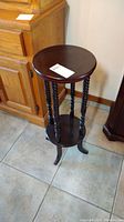 Full view of wooden plant stand with round top, twisted spindle legs, and three curved feet, positioned near wooden cabinet on tiled floor.