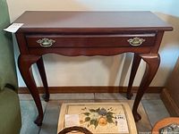 Full front view of the wooden side table showing the top surface, single drawer with brass handle, and curved legs.