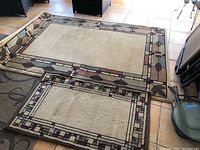 Pair of matching beige wool blend rugs with a cubist geometric border design, shown on tiled floor. Larger rug is rectangular and bigger than the smaller one placed on top of a different rug.