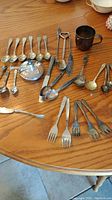 Overview of assortment of vintage spoons, forks, tongs, and a small silver plate cup on a wooden table.