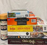 Stack of five 1000-piece puzzles and one board game on a white fabric background, showing puzzle themes and box details