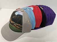 Eight baseball-style hats lined up side-by-side, showing various colors and the printed front panel with mountain golf course image.