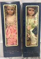Two porcelain dolls visible through clear window in navy blue boxes with gold trim, dressed in detailed outfits