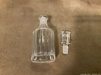 Clear glass faceted perfume bottle and matching stopper separated and placed on a surface, showing bottle shape and stopper details.