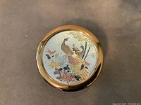 Top view close-up of round trinket dish lid featuring detailed peacock and floral imagery with gold plating.