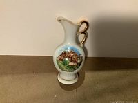 Front view of the porcelain miniature pitcher showing the scenic mountain design and gold accented handle.