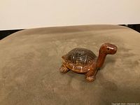 Photo of a small brown glazed pottery turtle figurine on a soft surface, showing body and shell detail.