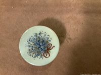 Top view of the round alabaster pill box lid with blue floral pattern and brown ribbon on a light background.