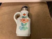 Milk glass syrup pitcher with metal lid and floral hand-painted decoration on front, sitting on brown surface.