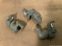Three grey resin elephant figurines pose assorted, lying on a beige surface showing white tusks and original yellowish paper stickers on undersides.