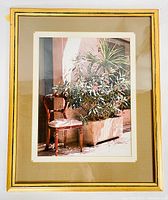 Framed color photograph showing wooden chair and planter with greenery and flowers against stucco wall.