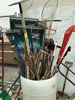 Bin full of assorted garden tools including rakes, shovels, hoes, and a fishing net, showing overall lot condition and contents