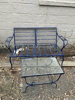 Blue iron two-seat outdoor chair with armrests next to a matching blue iron-framed glass-top table, set outdoors on a ground covered with dried leaves and dirt against a white wall.