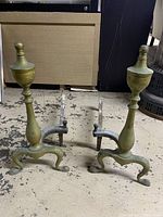 Pair of brass andirons standing on a mottled concrete floor showing the upright urn finials and tripod base legs.