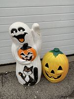 Two Halloween blow molds side by side outside on pavement in front of white garage door. White ghost holding pumpkin with cat and yellow pumpkin jack-o'-lantern.