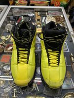 Front view of yellow and black Kobe Bryant basketball sneakers side by side on glass counter with various jewelry items beneath.