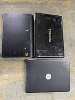 Top view of three laptops placed on a floor, showing HP, Toshiba, and Dell laptops with visible wear and scratches.