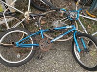 Blue Mongoose BMX bike with rusted chainring and missing right pedal lying on the ground among other bicycles.