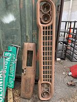 Photo showing two rusted metal car parts leaning against a green metal container, including a large grille with two circular headlight openings and a smaller panel with a rectangular cutout.