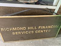 Front view of rectangular metal sign with gold raised letters 'RICHMOND HILL FINANCIAL SERVICES CENTER' on brown background with gold border.