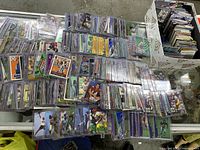 Top-down view of a large selection of 1990s sports cards in top loaders including rookies and inserts spread across a table and inside a box.