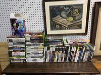 Wide shot showing all stacked video games from various consoles on a wooden surface with pegboard background and framed artwork.
