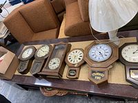 Six brown and dark wooden framed vintage wall clocks arranged on table showing various dial styles and pendulum boxes