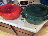 Two enameled cast iron baking dishes with lids on a countertop including a red smaller 13x9 inch and a green larger 16x11 inch dish.