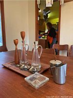 Overview of the set on wooden table showing wooden tray, two tall glass bottles with metal bases, two wooden candlesticks, metal creamer and sugar containers, glass covered dish in front.