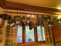 Wide shot of metal pots, pans, utensils, and strainers hanging from a wooden rack in front of a window with patterned curtains.