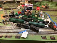 Wide shot of New Haven passenger train cars, assorted small train cars, and scenic modeling supplies including Woodland Scenics U-Mountain kit and several paint bottles on model railroad layout with control panel visible.