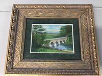 Framed landscape watercolour painting showing multi-arched stone bridge over river with surrounding trees, in ornate gold frame with dark green mat. Showing small wear marks on frame edges.