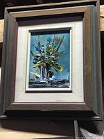 Framed oil painting showing a small floral arrangement in a vase with a blue background.